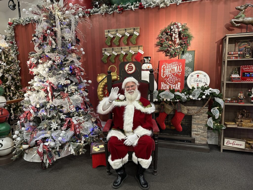 Santa Claus sitting in a festive chair, waving beside a decorated Christmas tree at the Santa Claus House in North Pole, Alaska.