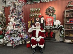 Santa Claus sitting in a festive chair, waving beside a decorated Christmas tree at the Santa Claus House in North Pole, Alaska.