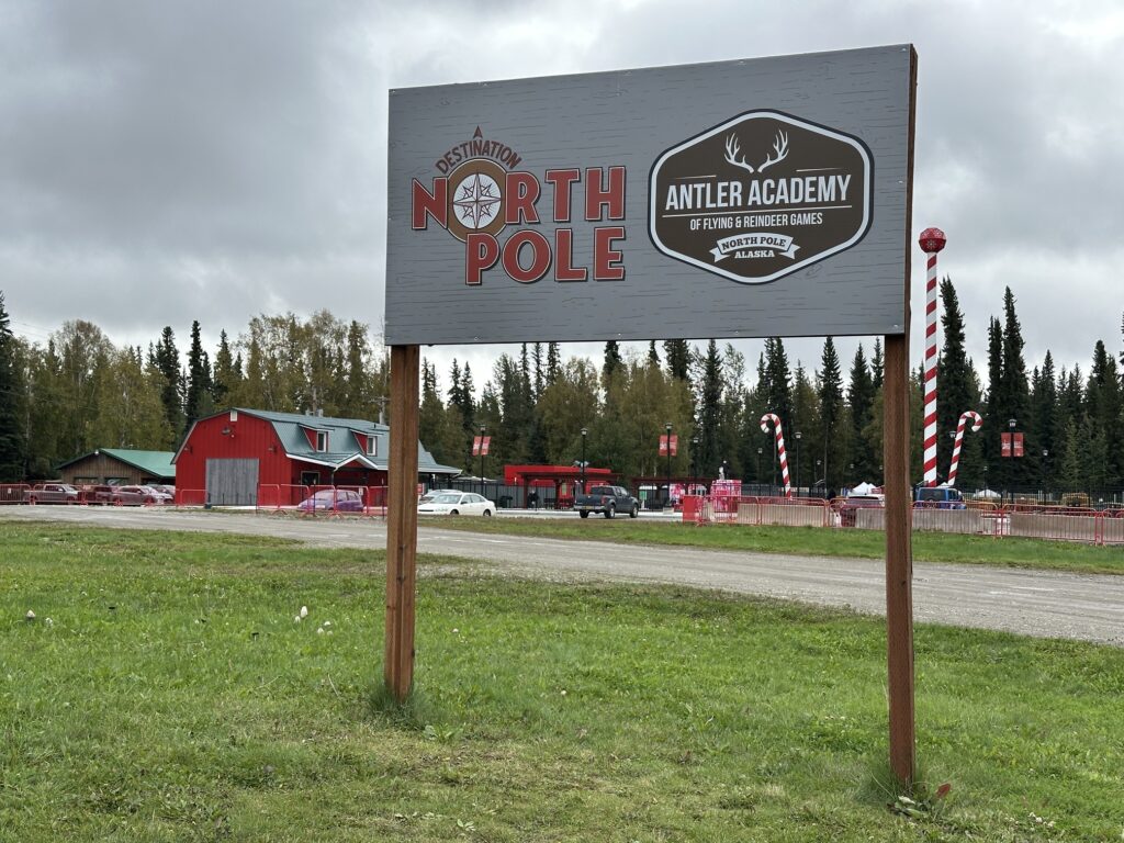Entrance sign for the Antler Academy of Flying & Reindeer Games at the North Pole in Alaska, with red barns and candy-cane light poles in the background.