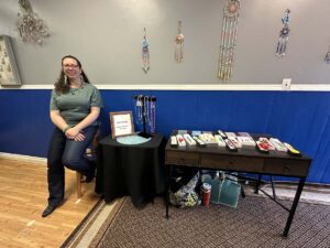 Artisan seated on a stool beside a display of handmade beaded jewellery—earrings, bracelets, and necklaces—inside an indoor vendor booth with dreamcatchers on a grey-and-blue wall.