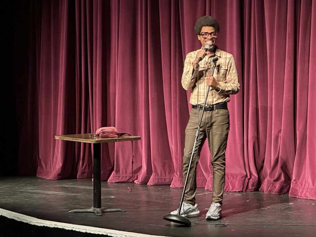 Comedian Mike E. Winfield performing on stage in Fairbanks, Alaska, with a microphone in hand and a red curtain backdrop.