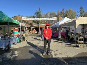Executive Director/ Farmer standing in front of the Tanana Valley Farmers Market with vendor tents around.