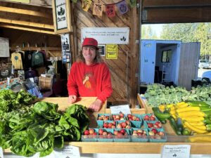 Smiling vendor inside a produce stall with lettuces, cucumbers, squash, and apples.