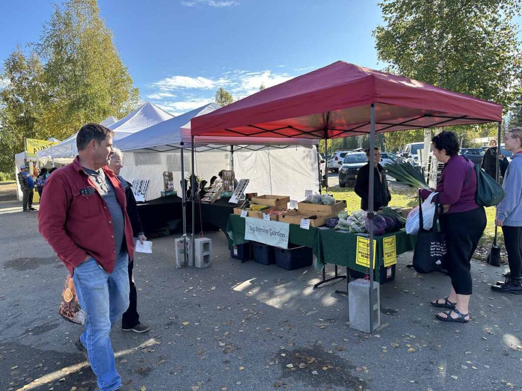 Shoppers chatting with a farmer under a red canopy at an outdoor market.