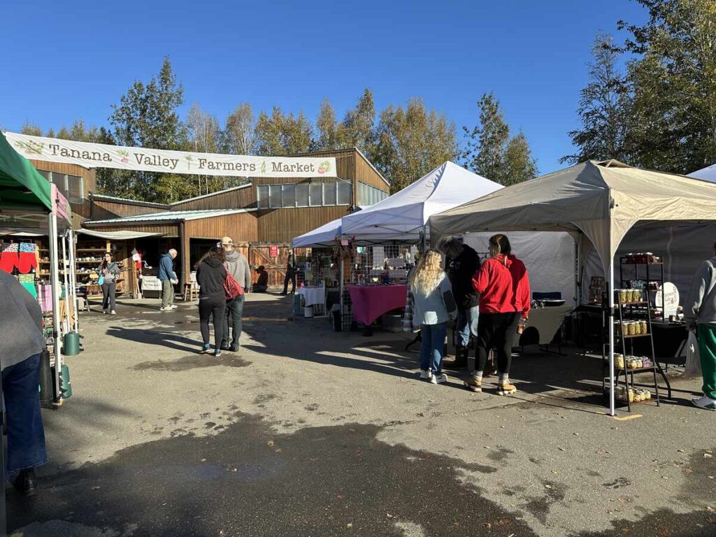 Shoppers browse vendor tents in front of the wooden Tanana Valley Farmers Market building, a banner reading “Tanana Valley Farmers Market” hanging overhead on a clear morning.