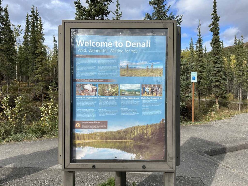 Information kiosk titled “Welcome to Denali” on a paved path, bordered by spruce and early-autumn foliage.
