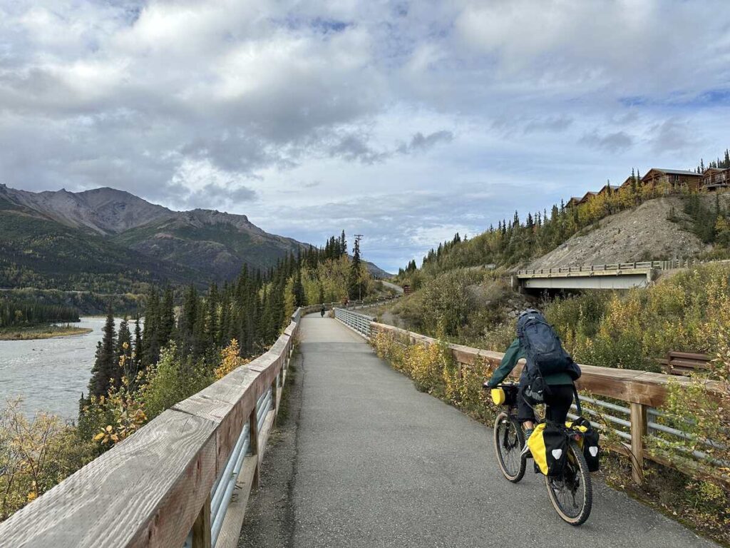 Touring cyclist with panniers riding a paved riverside path toward mountain foothills; cabins dot a hillside to the right.