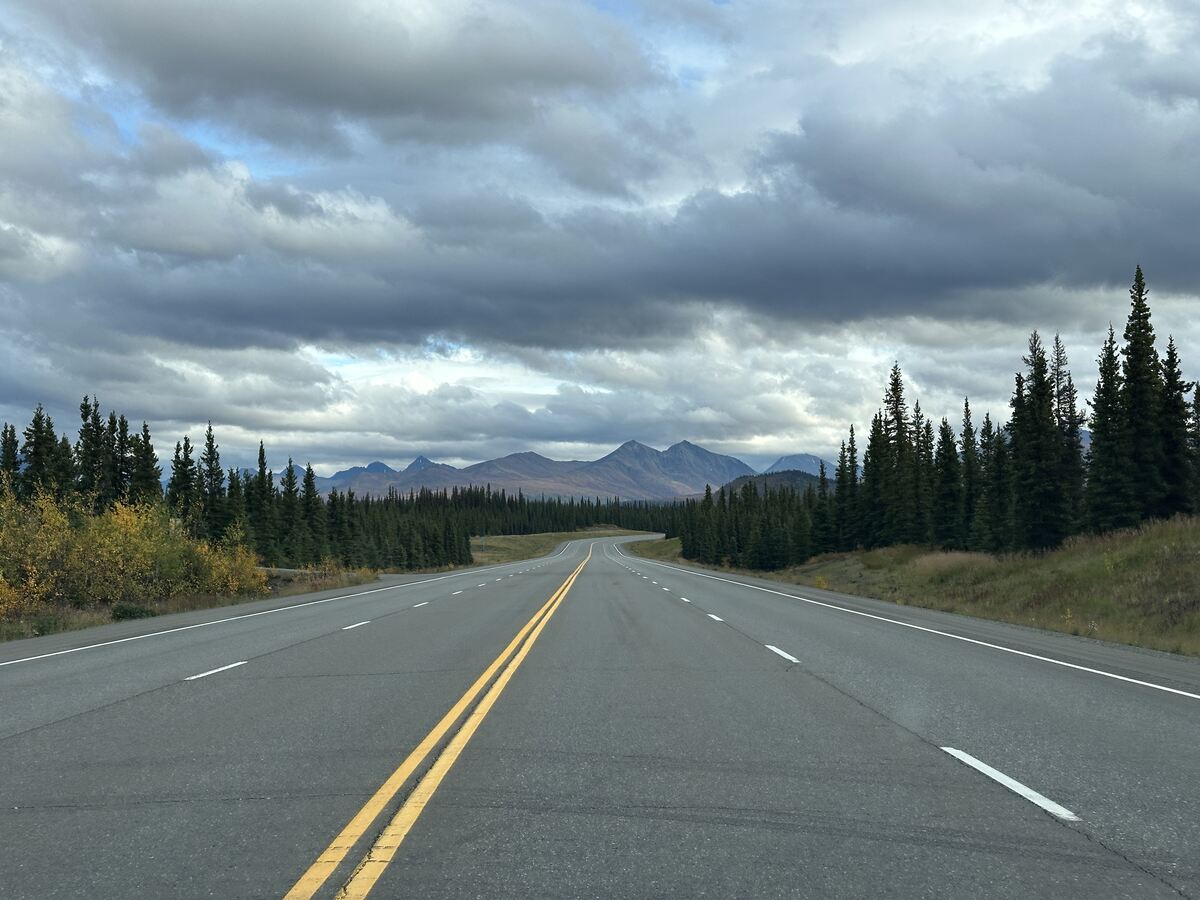 Empty two-lane highway through spruce forest toward distant mountains beneath layered grey clouds.