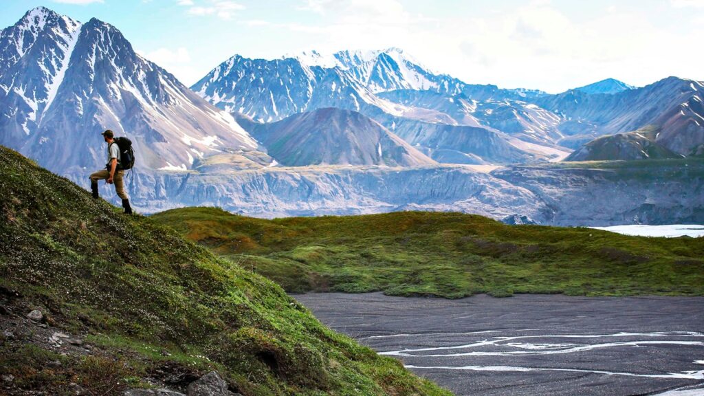 Hiker traverses a green hillside above a braided river with steep, grey mountains beyond.