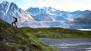 Hiker traverses a green hillside above a braided river with steep, grey mountains beyond.