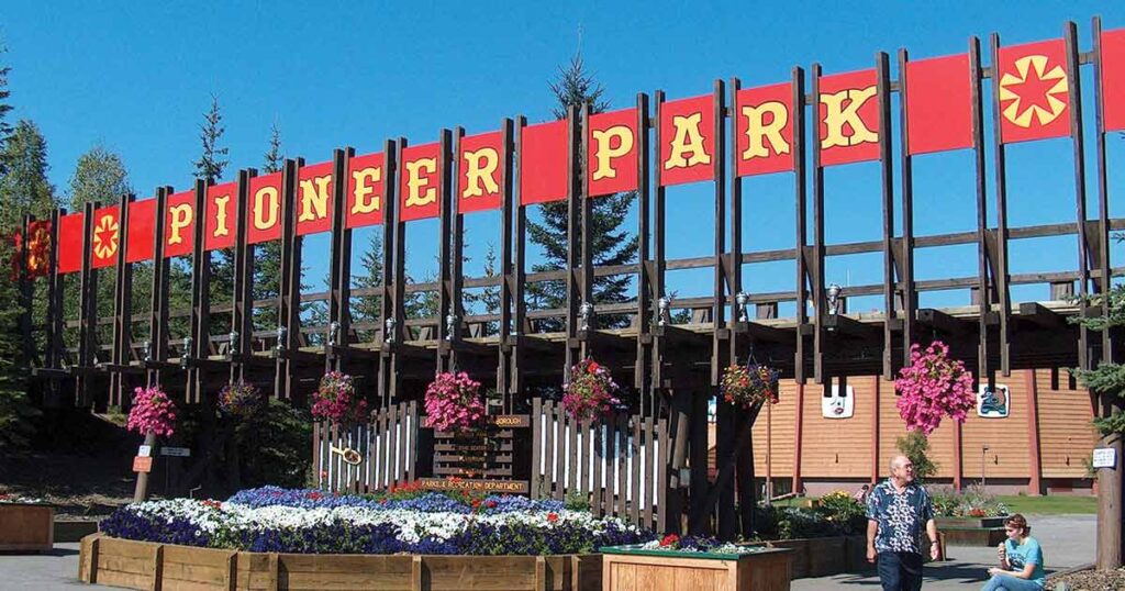 Entrance sign at Pioneer Park in Fairbanks, Alaska, with bold red-and-gold lettering and hanging flower baskets, marking the gateway to the historic theme park.