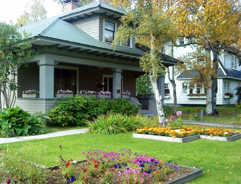 Exterior of the Alaska Heritage House in Fairbanks, Alaska, showing its historic architecture with a wide porch, flower boxes, and landscaped gardens with blooming flowers in front.