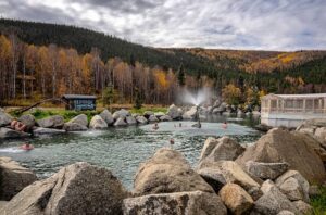 Visitors relax in the outdoor natural rock pool at Chena Hot Springs Resort, surrounded by golden autumn trees and forested hills.