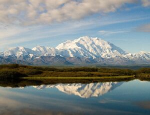Denali’s snow-clad massif reflected in a still pond under broken clouds.