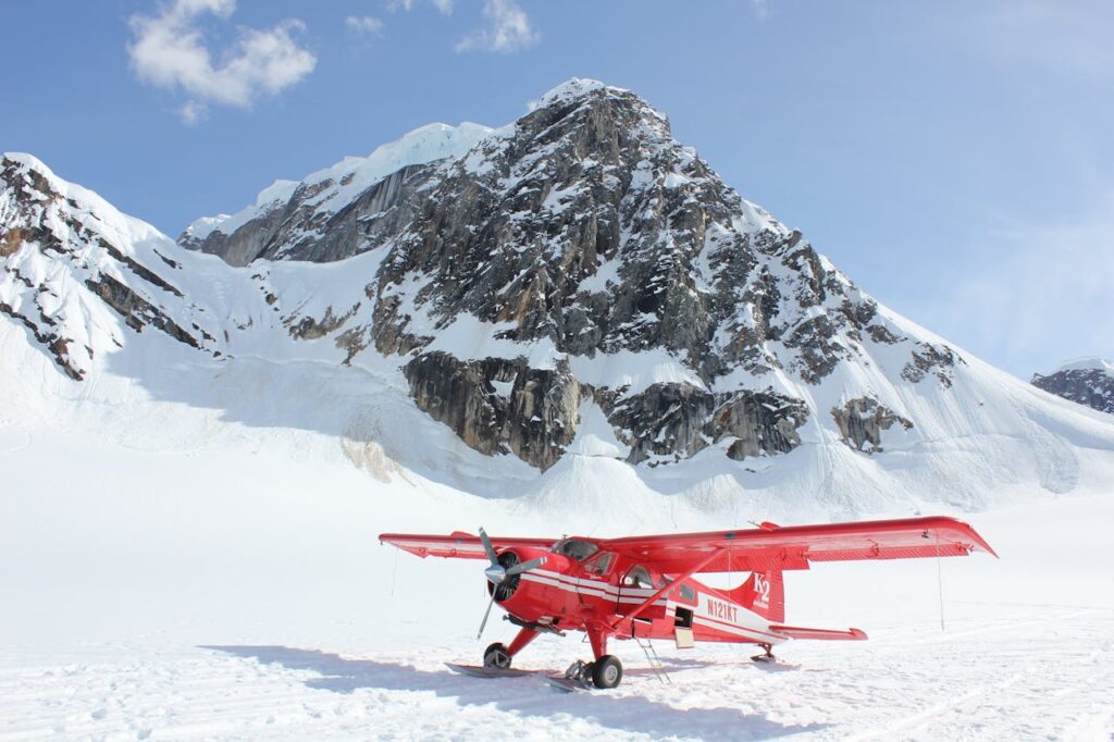 Red ski-plane parked on a sunlit glacier beneath a steep, rocky, snow-covered peak.