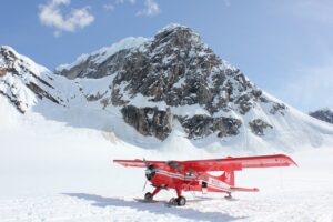 Red ski-plane parked on a sunlit glacier beneath a steep, rocky, snow-covered peak.