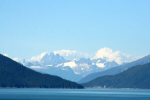 Snow-capped, jagged peaks and glacier tongues rise beyond a forested fjord on a clear summer day in Alaska.