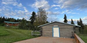 A rustic log cabin with a white door and blue trim, labeled “The Night Watch,” sits on a wooden deck at A Taste of Alaska Lodge. Behind it, the main lodge is visible under a bright blue sky dotted with clouds, surrounded by tall evergreens and lush grass.