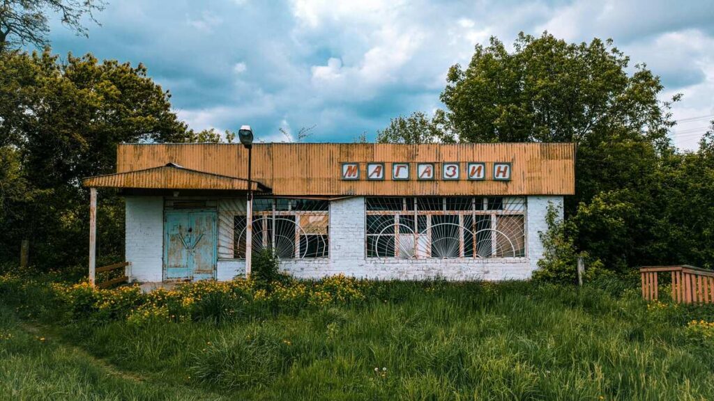 An abandoned village shop with broken windows and overgrown grass, its faded Cyrillic sign hinting at lives and economies left behind.