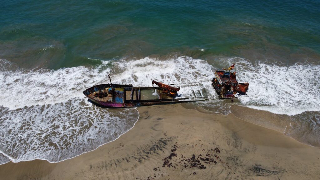 A rusted shipwreck lies broken on the shoreline as waves crash around it, a stark metaphor for human excess and discard.