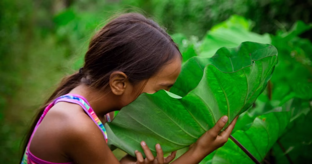 A young child with long dark hair leans in close to a huge bright green leaf, holding it gently with both hands and pressing her face toward it as if smelling or listening. She is surrounded by lush plants, and the background is soft and green.
