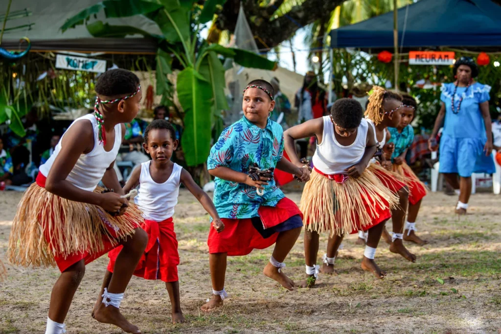 A group of children dance barefoot outdoors on sandy ground. They are dressed in bright clothing — red skirts or shorts, some with woven grass skirts and white tank tops, others in patterned shirts and headbands. Their knees are bent and their hands are poised in motion, focused and energetic. Adults and palm leaves can be seen in the background, suggesting a community celebration.