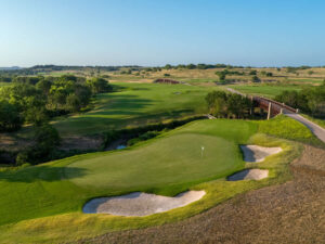 Aerial view of the 18th hole at Fields Ranch East, with a bright green putting surface framed by bunkers, a tree-lined fairway stretching into the distance and a rust-coloured bridge crossing a small creek under a clear blue Texas sky.
