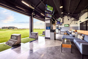 Open-air golf hitting bays at Ice House, featuring turf tee decks facing the driving range, plush lounge seating, and multiple TV screens hanging from an industrial-style ceiling.