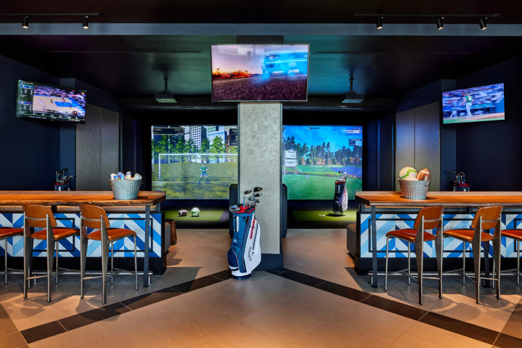 Indoor lounge with bar-height tables and leather stools facing two large Topgolf Swing Suite simulator screens, surrounded by multiple TVs showing different sports.