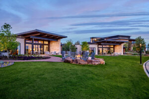 Evening view of low-slung Ranch House buildings with large windows, surrounded by manicured lawn and landscaping, and a circular firepit ringed with rocking chairs.