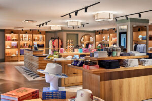 Bright resort boutique with wooden display tables and shelves showcasing hats, books, resort wear, accessories and colourful gifts under soft ceiling lights.