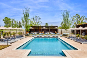 Rectangular pool surrounded by cushioned loungers, umbrellas and minimalist cabanas, framed by young trees and blue sky.