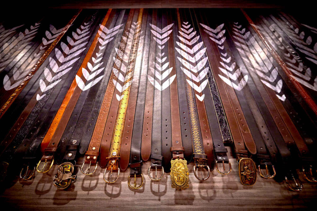 Art installation made from rows of vintage leather belts in shades of brown and black, laid side by side with ornate metal buckles at the bottom and white arrow or feather motifs painted across the straps.