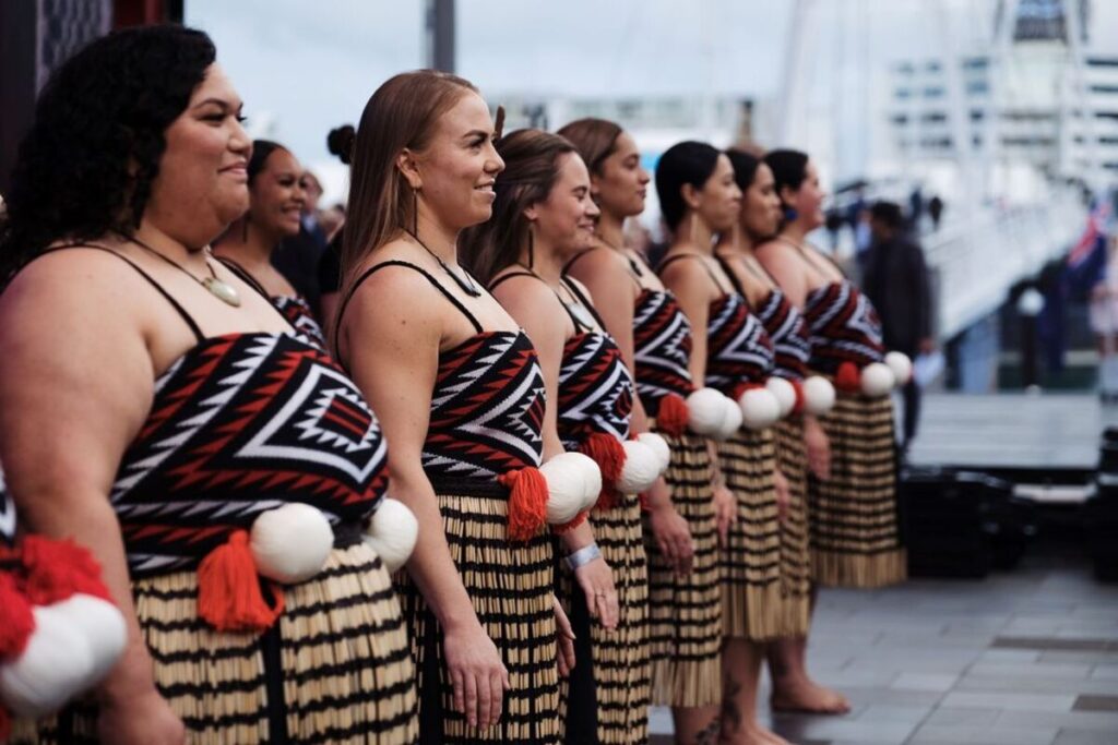 A line of Māori women stands side by side outdoors, wearing traditional clothing. Their outfits include patterned black, red, and white bodices and flax skirts with tassels, along with poi balls tied at the waist. They face forward with calm focus, preparing to perform. The background is slightly blurred with people and harbour structures.