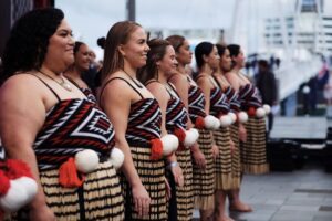 A line of Māori women stands side by side outdoors, wearing traditional clothing. Their outfits include patterned black, red, and white bodices and flax skirts with tassels, along with poi balls tied at the waist. They face forward with calm focus, preparing to perform. The background is slightly blurred with people and harbour structures.