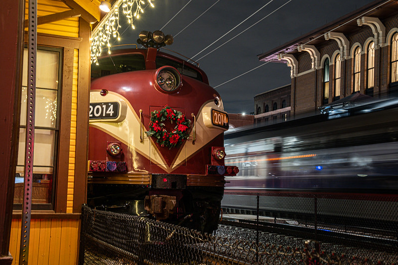 A vintage red locomotive numbered 2014 is parked beside a yellow wooden depot, its front decorated with a Christmas wreath and framed by twinkling white lights hanging from the roofline. Overhead wires stretch across the night sky, and in the background a second train streaks past in a blur, highlighting the stillness of the old engine against the motion of modern rail.