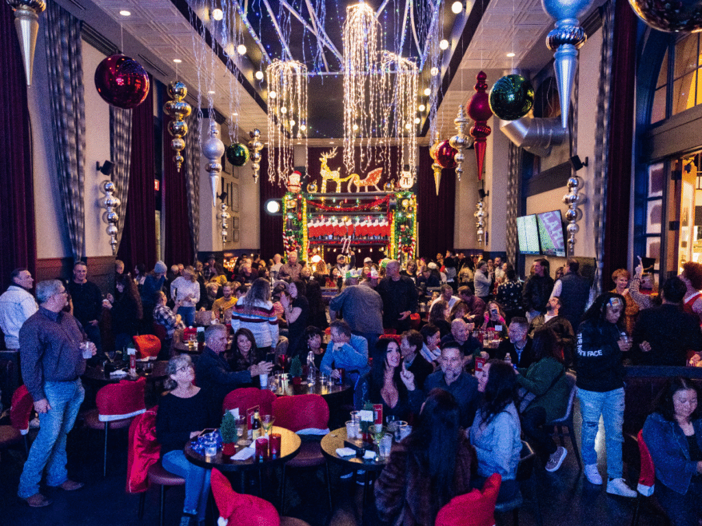 A packed holiday bar with people seated at small tables, chatting and taking photos beneath dramatic ceiling garlands, hanging ornaments, and twinkling string lights framing a brightly decorated stage.