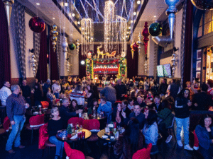A packed holiday bar with people seated at small tables, chatting and taking photos beneath dramatic ceiling garlands, hanging ornaments, and twinkling string lights framing a brightly decorated stage.