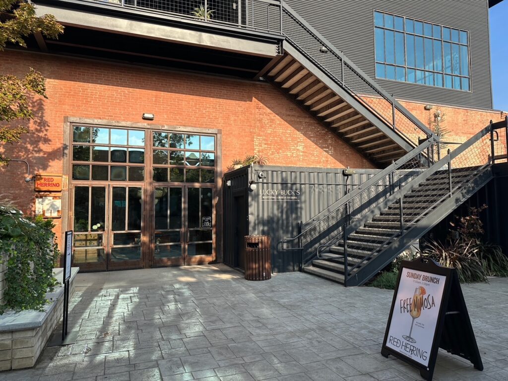 A brick courtyard at Hotel Herringbone with the tall glass doors of Red Herring restaurant on the left and a dark shipping container labelled “Lucky Buck’s” tucked beneath an exterior staircase on the right. Metal stairs rise to an upper level, and a sandwich board in the foreground advertises Sunday brunch with a “Free Mimosa” sign. Potted plants and low greenery soften the edges of the space.