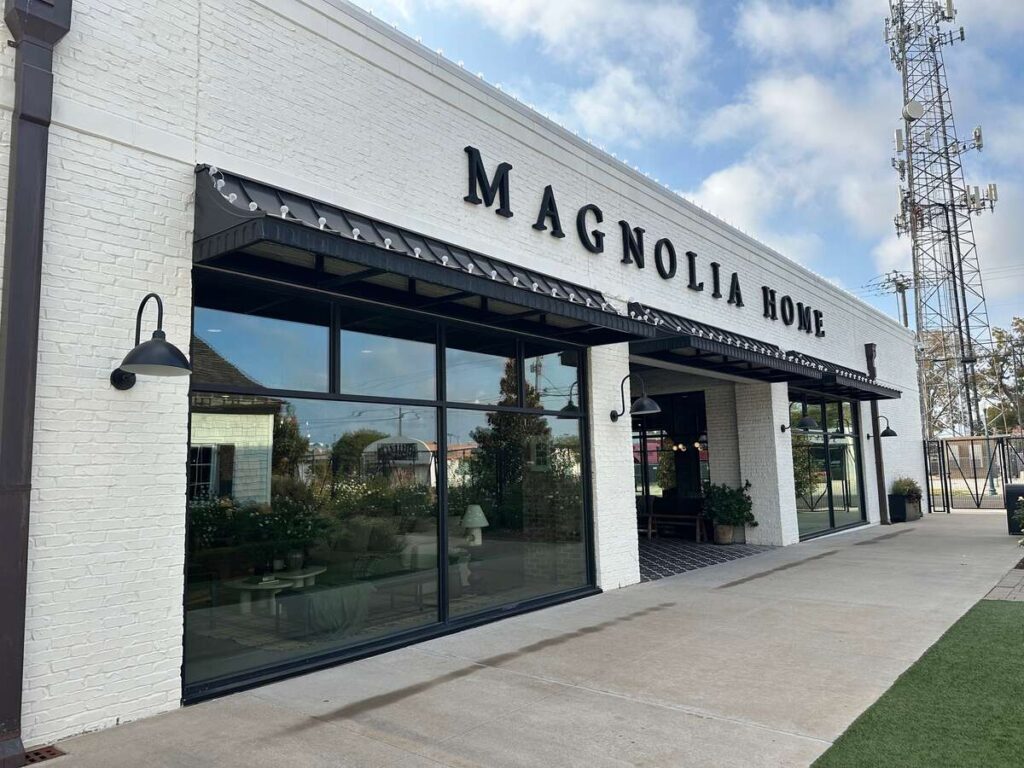 A low, white-brick building with black awnings and large glass windows stretches along a clean sidewalk. The sign “Magnolia Home” spans the top in simple black letters. Through the windows, soft furnishings and greenery are faintly visible. A cell tower and blue sky with scattered clouds rise behind the building.