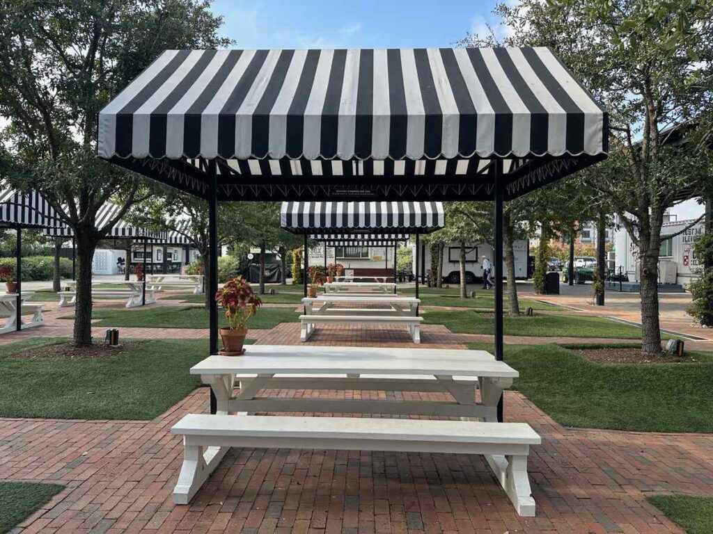 A row of white picnic tables stretches down a brick pathway, each covered by a black-and-white striped canopy. Neatly trimmed artificial turf fills the spaces between the bricks, and young trees frame the scene. A small potted plant sits on the nearest table, and the whole area is empty, waiting for visitors.