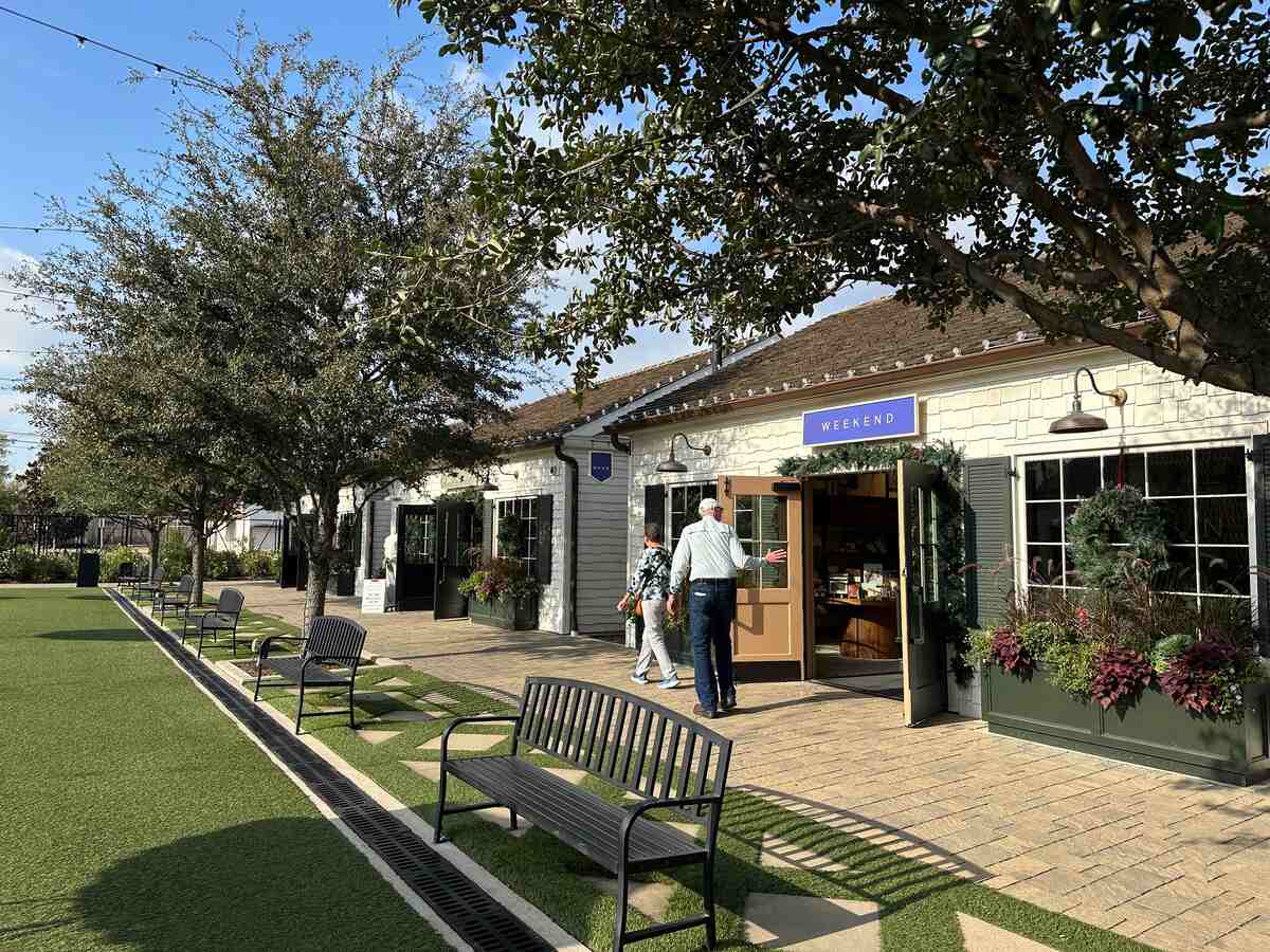 A row of cottage-style shopfronts with green shutters and window boxes faces a long strip of artificial turf dotted with metal benches. One shop, with a purple “Weekend” sign over the door, is open; a couple stands in the doorway talking. Mature trees cast dappled shade across the walkway.