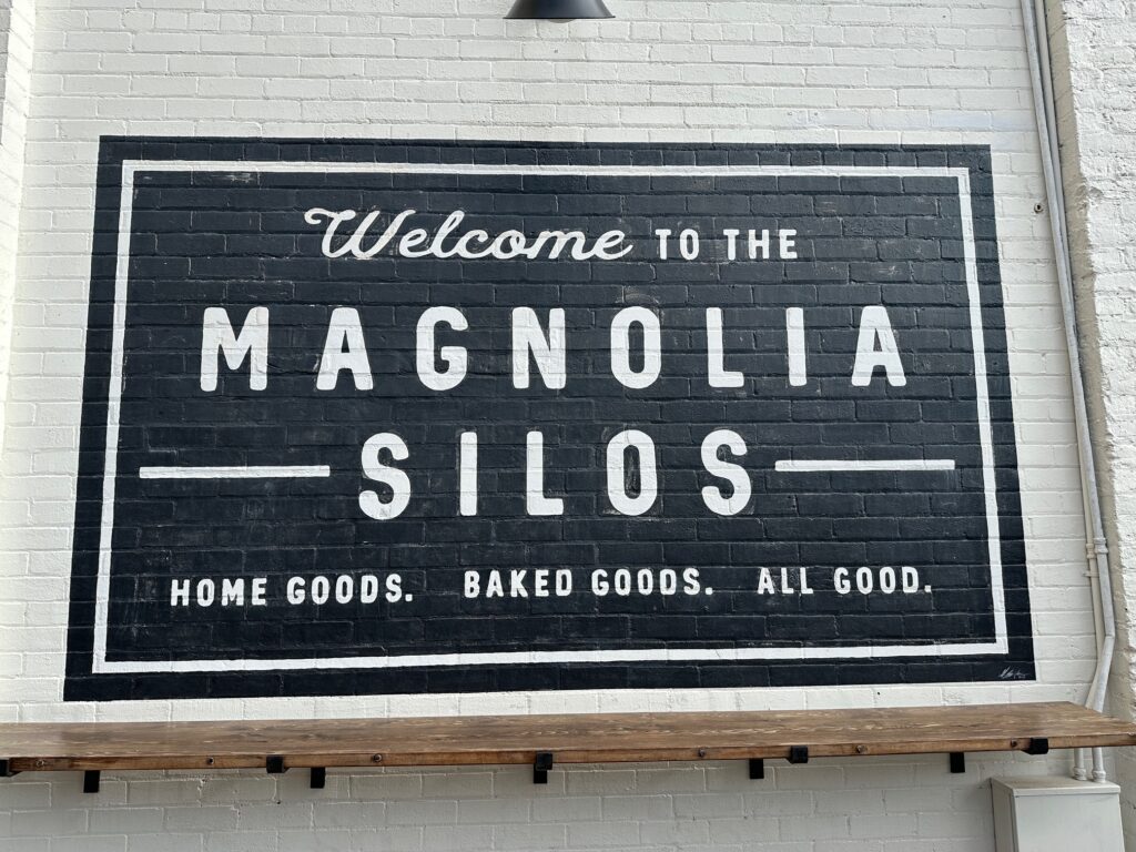 A black-and-white painted sign fills a section of white brick wall. The words read “Welcome to the Magnolia Silos – Home Goods. Baked Goods. All Good.” in simple lettering, framed by a white border. A narrow wooden ledge runs along the bottom of the wall.