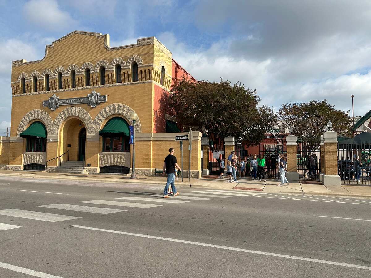 A mustard-yellow, Romanesque-style building with arched windows and stone trim stands on a corner across from a crosswalk. A sign above the door reads “Dr Pepper Museum.” To the right, an iron gate leads into a courtyard where people are lined up to enter. The sky is partly cloudy, and a few pedestrians cross the street in front.