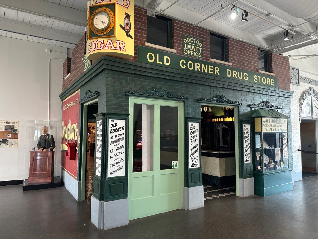 Inside the museum, a full-scale recreation of the Old Corner Drug Store features green and red brick walls, vintage painted signs, and a glass display window. The entrance is open, revealing a marble soda fountain counter inside. To the left, a life-sized mannequin of a suited man stands behind a small lectern, as if addressing visitors.
