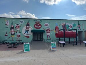A wide, turquoise wall is covered with a playful mural of cartoon Dr Pepper bottles and cups with arms and legs, dancing and holding signs like “More Fun!” and “More Exhibits!” In front of the mural sits a large red industrial tank on metal supports, with interpretive plaques. Picnic tables and two green bilingual “Soda Fountain” signs stand on the concrete plaza under a bright blue sky.