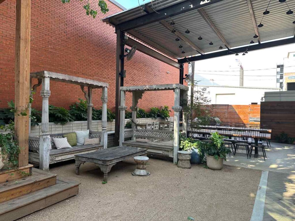 A cosy outdoor courtyard with a gravel floor features two long, carved wooden daybeds under simple four-post frames, each piled with soft cushions in muted colours. A weathered wooden coffee table and a small round stool sit between them. Overhead, a metal roof with string lights provides shade. Lush green plants line the low brick planter along a tall red-brick wall, and in the background a long wooden table with slim black chairs offers extra seating.