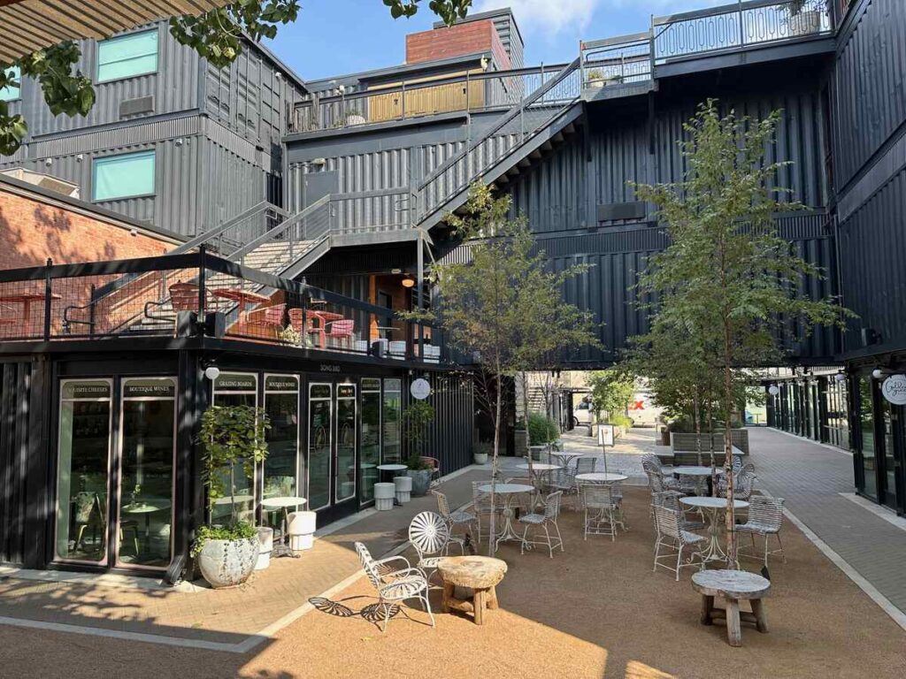 A cosy outdoor courtyard with a gravel floor features two long, carved wooden daybeds under simple four-post frames, each piled with soft cushions in muted colours. A weathered wooden coffee table and a small round stool sit between them. Overhead, a metal roof with string lights provides shade. Lush green plants line the low brick planter along a tall red-brick wall, and in the background a long wooden table with slim black chairs offers extra seating.