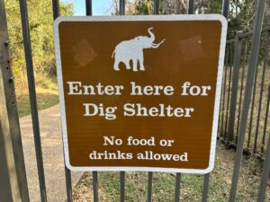 Close-up of a brown metal sign mounted on a black fence, with a white silhouette of a mammoth at the top and white text reading “Enter here for Dig Shelter – No food or drinks allowed.” Behind the sign, a paved path curves into a wooded area of green shrubs and trees.