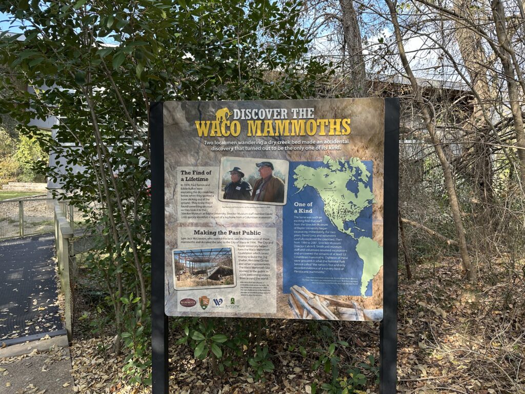 An outdoor interpretive sign titled “Discover the Waco Mammoths” stands beside a wooded path, with photos of the original discoverers, a map of North America, and text explaining how the site was found.